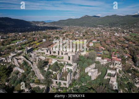 Una vista aerea a grandangolo cattura le antiche rovine di pietra della città fantasma di Kayakoy circondata da vegetazione verde, con il moderno villaggio e lontano h Foto Stock