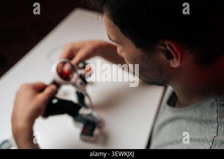 Vista sopra la spalla di un giovane uomo concentrato nell'assemblare e riparare un piccolo gadget elettronico. Una bassa profondità di campo sfoca le mani e il dispositivo Foto Stock