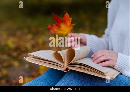 Primo piano di una donna in maglione seduta all'aperto con foglie autunnali colorate e un libro aperto con pagine piegate a forma di cuore che simboleggiano l'amore per i readi Foto Stock