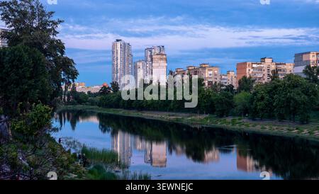 Panorama urbano che mostra alti edifici moderni e vecchi edifici residenziali lungo un fiume sereno, con riflessi urbani che appaiono sullo sfondo Foto Stock