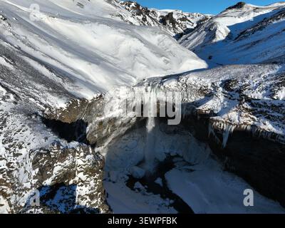 Vista aerea della cascata di Kvernufoss che scende lungo un canyon roccioso innevato con formazioni di ghiaccio e aspre scogliere a Evindarholar, Rangarthing eyst Foto Stock