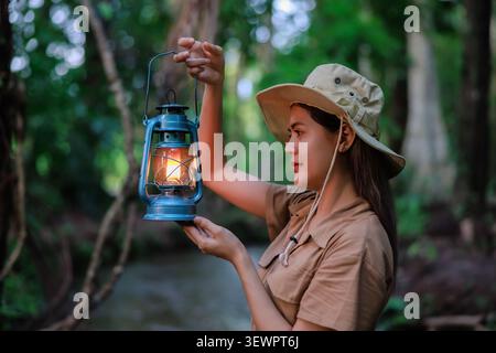 Una donna in abbigliamento outdoor esplora una foresta mentre porta con sé una lanterna luminosa che simboleggia l'avventura alla scoperta e lo spirito di esplorazione. Foto Stock