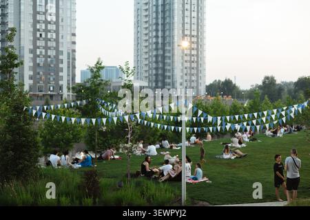 Kiev, Ucraina - 27 giugno 2026 i residenti e i visitatori si rilassano su un prato verde in un moderno quartiere residenziale durante una calda serata estiva, godendosi Foto Stock
