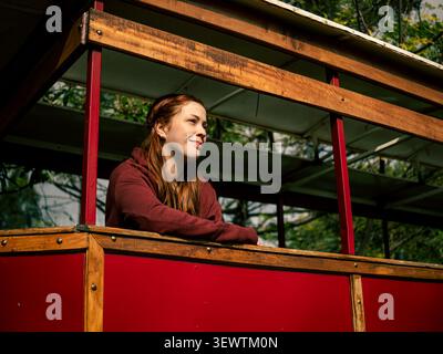 La donna si appoggia su una ringhiera rossa in legno. Ha lunghi capelli castani e indossa una felpa con cappuccio marrone. Sorride dolcemente mentre guarda di lato. La t Foto Stock
