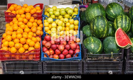 Frutta fresca assortita in casse presso un bancone del mercato, con arance, mele verdi, mele rosse e angurie, con un anguria a fette visibile Foto Stock