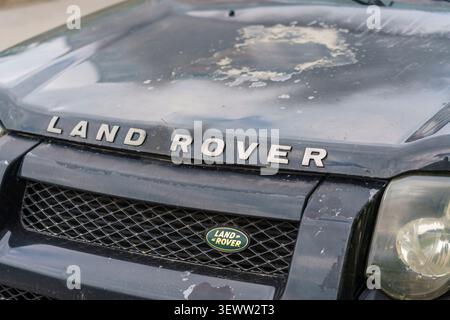 Vista ravvicinata della cappa nera del SUV Land Rover con graffi e segni di usura visibili, con la griglia anteriore a nido d'ape, la scritta cromata e il logo ovale Foto Stock