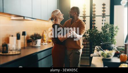Coppia felice, danzante e matura in cucina per cucinare, legare e amare musica, canzoni e preparazioni per i pasti. Interrazziale, matrimonio e anziano e.. Foto Stock