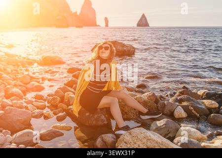 Il tramonto sulla costa delle donne si siede su una spiaggia rocciosa indossando una giacca gialla e occhiali da sole, godendosi una serena serata sulla riva Foto Stock