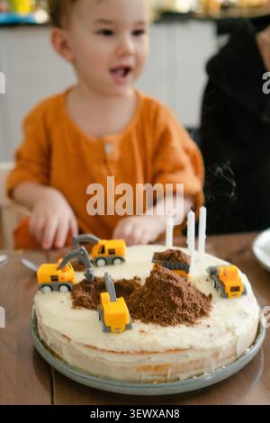 il bambino sorride dopo aver soffiato le candele sulla sua torta di compleanno. il bambino ha tre anni. la torta fatta in casa è decorata come un cantiere con camion giocattolo. festa di famiglia Foto Stock