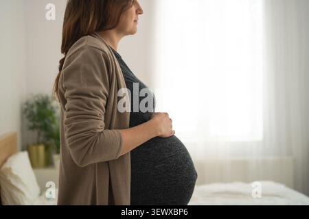 Aspettatevi una madre in piedi con un batuffolo visibile in un'accogliente camera da letto con luce naturale. La futura mamma irradia felicità accanto al letto con decorazioni vegetali. Donna incinta che gode di calma e felice attesa a casa Foto Stock