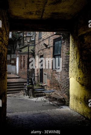 Una vista incorniciata che guarda attraverso un arco di pietra scuro in un vicolo tranquillo e soleggiato a Durham, caratterizzato da un lampione vintage e una bicicletta solitaria. Foto Stock
