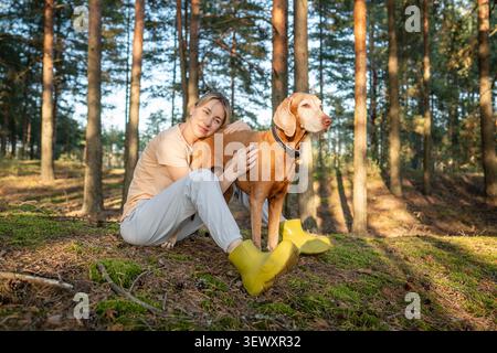 Donna felice e rilassata proprietaria di animali domestici comodamente seduta sul prato della foresta che posa la testa sul cane Vizsla. Foto Stock
