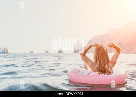 Donna, rilassante, oceano. Donna bionda che galleggia su un anello gonfiabile di ciambella rosa in mare aperto vicino alle scogliere durante le vacanze estive con spazio per copiare. Foto Stock