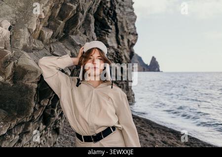 Donna ushanka costiera che indossa un elegante cappello invernale si appoggia su una scogliera rocciosa vicino al mare in una giornata nuvolosa Foto Stock