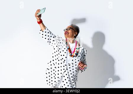 Donna anziana nera che indossa un ritratto di abbigliamento moderno sullo sfondo - donna anziana con capelli corti grigi e occhiali che fa un selfie con il suo smartphone, Foto Stock