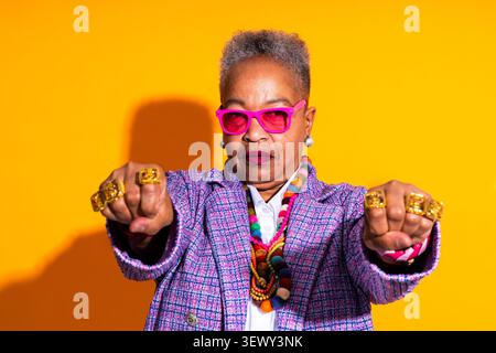 Donna anziana nera che indossa un ritratto di abbigliamento moderno sullo sfondo - donna afroamericana anziana che posa con un atteggiamento sicuro. Indossa occhiali rosa Foto Stock
