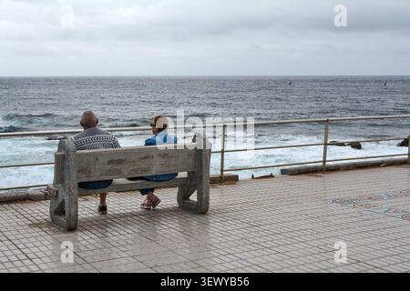Agaete, Spagna - 29 giugno 2026: Un uomo e una donna siedono su una panchina di cemento lungo una passeggiata costiera ad Agaete, Spagna, guardando l'oceano blu ondulato Foto Stock