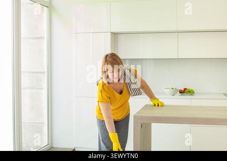 Una donna che indossa guanti gialli sta pulendo un tavolo da cucina in una stanza luminosa. Utilizza un panno per pulire la superficie stando vicino a una finestra con il sole Foto Stock