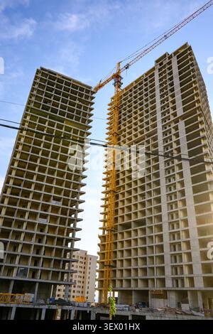 Alti e torreggianti edifici in costruzione. Una grande gru solleva i materiali tra le strutture. Cielo blu chiaro con luce solare soffusa. Sviluppo urbano Foto Stock