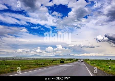 Lungo rettilineo si estende verso montagne lontane. Cielo pieno di soffici nuvole bianche e macchie blu intenso Foto Stock