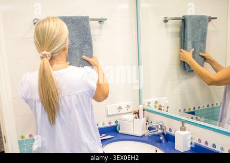 Un'individuo grazioso appende con cura un asciugamano morbido su una rastrelliera d'argento in un bagno vivace. L'atmosfera serena riflette una bellezza semplice e mattine tranquille, arricchendo i rituali quotidiani. Foto Stock