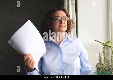 Donna di mezza età che tiene in mano un documento e guarda premurosa davanti a una finestra in ufficio Foto Stock