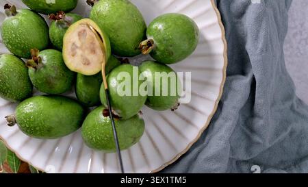 Una foto in alto di vivaci frutti verdi feijoa, una tagliata a metà per rivelare l'interno cremoso, disposta su un piatto bianco testurizzato con un elegante cucchiaio Foto Stock
