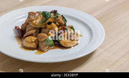 Un piatto bianco di tre tazze di pollo con funghi e foglie di basilico si trova su un tavolo di legno chiaro. La salsa di soia marrone dorato comprende teneri pezzi di pollo insieme a funghi ed erbe aromatiche in questo classico piatto cinese cotto in padella. Foto Stock