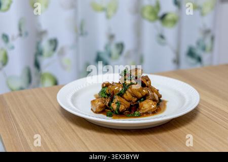 Un piatto di tre tazze di pollo si siede su un tavolo da pranzo in legno, i pezzi di pollo brasati glassati in ricca salsa di soia e sesamo con profumate foglie di basilico e peperoncino secco. Un classico piatto cinese casalingo servito a cena di famiglia. Foto Stock