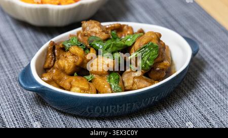 Tre tazze di pollo con basilico fresco e glassa di soia lucida sono servite in un piatto ovale blu in ceramica su un tavolo di legno. Sullo sfondo si trova una ciotola di pomodoro e uova fritte, che completa l'ambiente dei piatti cinesi fatti in casa. Foto Stock