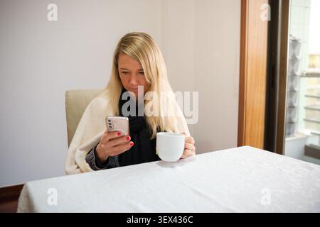 Giovane donna seduta a un tavolo a controllare il telefono e a bere una bevanda Foto Stock