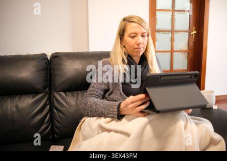 Donna seduta su un divano, coperta da una coperta, che utilizza un tablet a casa Foto Stock