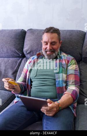 Uomo sorridente mentre acquisti comodamente online a casa, utilizzando una carta di credito e un tablet digitale per un pagamento sicuro Foto Stock