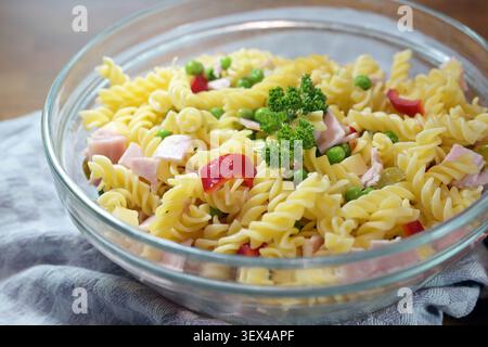 Insalata di pasta colorata con fusilli, peperoni, piselli, prosciutto cotto e prezzemolo guarnire in una ciotola di vetro su un asciugamano da cucina grigio blu, serba per feste fatte in casa Foto Stock
