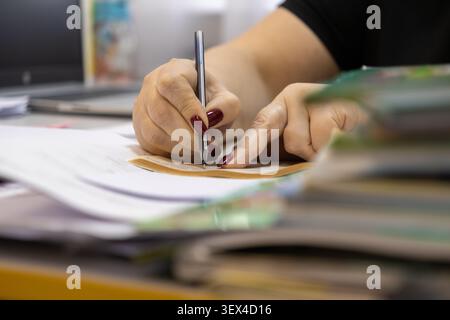 Primo piano di una mano adulta che firma documenti in una postazione disordinata, che rappresenta diligenza, attenzione ai dettagli e produttività professionale organizzata Foto Stock