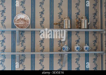 Ripiani bianchi a parete con pentole di caffè vintage in metallo, tazze con motivi blu e un piatto decorativo su carta da parati floreale a righe in un'accogliente cucitura rustica Foto Stock