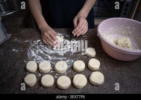 Le mani della persona modellano e porgono con cura l'impasto in piccoli pezzi circolari su un bancone da cucina spolverato di farina, preparando gli ingredienti per la cottura Foto Stock