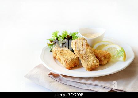 Bastoncini di pesce impanati croccanti serviti con insalata verde fresca, spicchi di limone e maionese su un piatto bianco, sfondo bianco. Copia spazio. Foto Stock