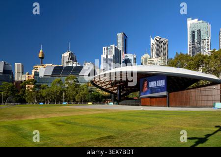 Australia, nuovo Galles del Sud, Sydney, Darling Harbour, Tumbalong Park, prato e palcoscenico coperto nel parco con lo skyline del centro città sullo sfondo, tra cui la Sydney Tower e gli edifici del CBD Foto Stock