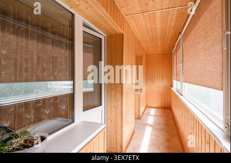 Balcone rivestito in legno con finestre e tende a rullo marroni. La luce splende sul pavimento piastrellato. Una pianta siede sul davanzale della finestra Foto Stock