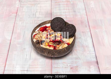 Parfait di yogurt condito con biscotti Oreo, frutti di bosco, muesli e fette di banana servito in una ciotola di cocco su legno bianco. Foto Stock