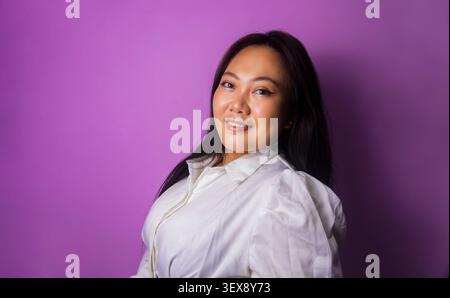 Giovane donna asiatica in affari con una camicetta bianca sorride mentre posa su uno sfondo studio viola Foto Stock