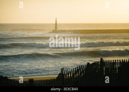 Tramonto dorato sull'Oceano Atlantico con onde ondulate, un faro frangiflutti e una recinzione di dune intempestiva che incornicia una costa costiera spazzata dal vento. Foto Stock