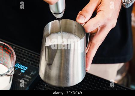 Barista esperto che utilizza un pannarello elettrico portatile che filtra latte fresco in caraffa in acciaio inox per creare schiuma per la preparazione di latte o cappuccino Foto Stock