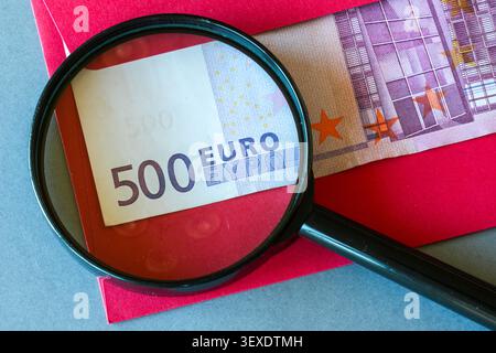 La lente d'ingrandimento rivela una busta rossa nascosta da 500 euro che simboleggia il concetto di pagamento segreto e ricchezza Foto Stock
