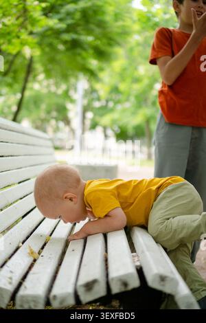 Allegro fratello maggiore che applaude mentre il bambino giace sulla panchina bianca all'aperto. Il bambino che riposa felicemente mentre il fratello festeggia con entusiasmo. Due bambini che condividono momenti felici e giocosi nella natura Foto Stock