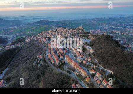 Vista aerea della storica Torre Guaita e dei circostanti edifici medievali in pietra, strade tortuose e lussureggiante paesaggio montano al tramonto nella città d'Oro Foto Stock
