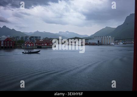 Pittoresco villaggio di pescatori norvegese a Lofoten con edifici tradizionali rossi, calme acque del porto, spettacolare sfondo di montagna e ormeggiato Foto Stock