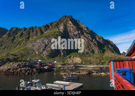 Pittoresco villaggio di pescatori norvegese annidato sotto vette torreggianti nelle isole Lofoten. Le tradizionali cabine rosse punteggiano le coste rocciose con ormeggio Foto Stock