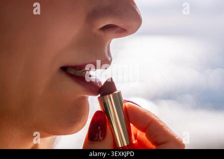 Applicazione di trucco rossetto donna applica il balsamo cosmetico per labbra alla bocca di fronte allo spazio di copia bokeh dell'acqua oceanica illuminata dal sole Foto Stock